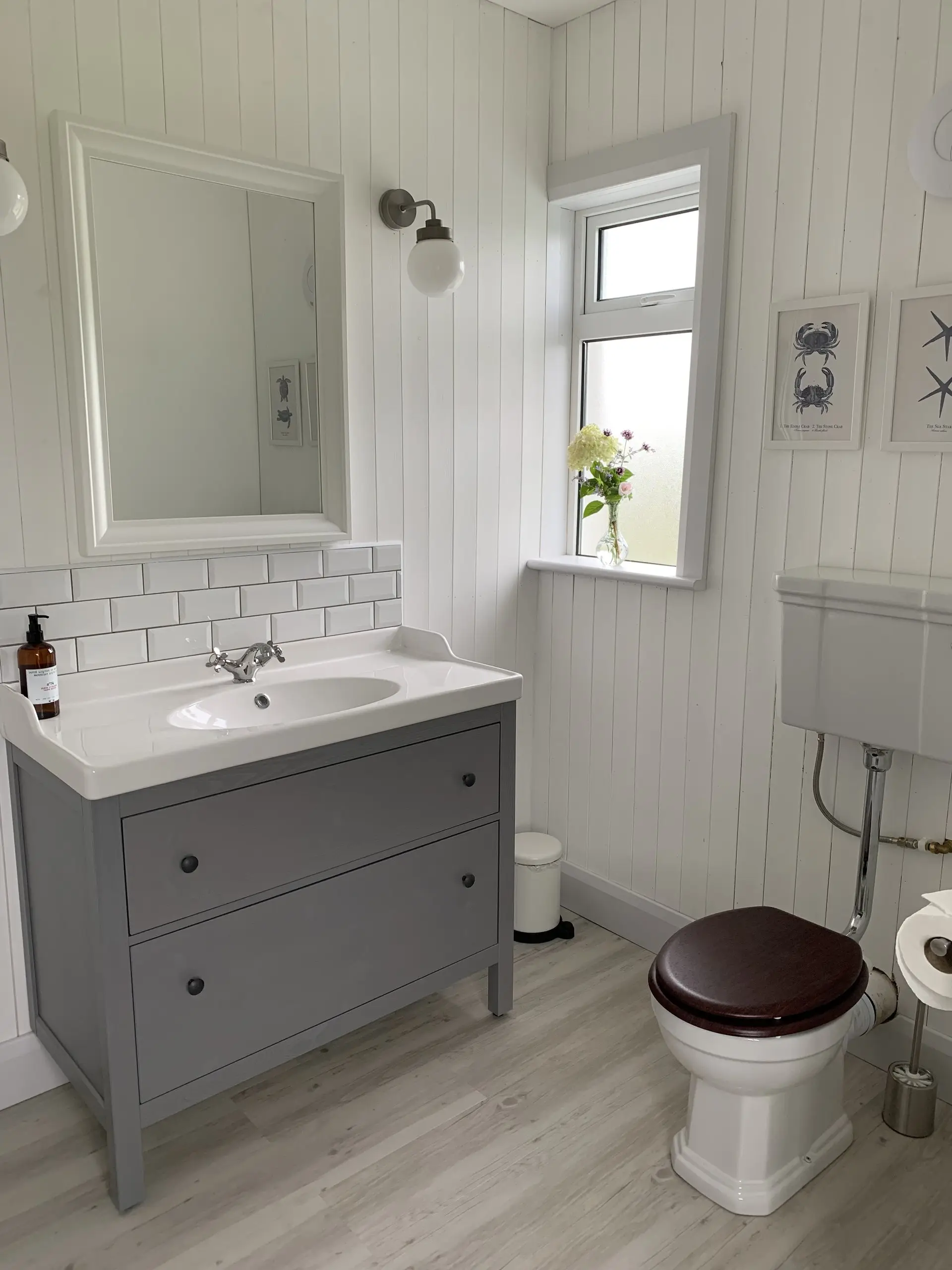 A bathroom vanity: a grey unit with a basin, white metro tiling and a window above.