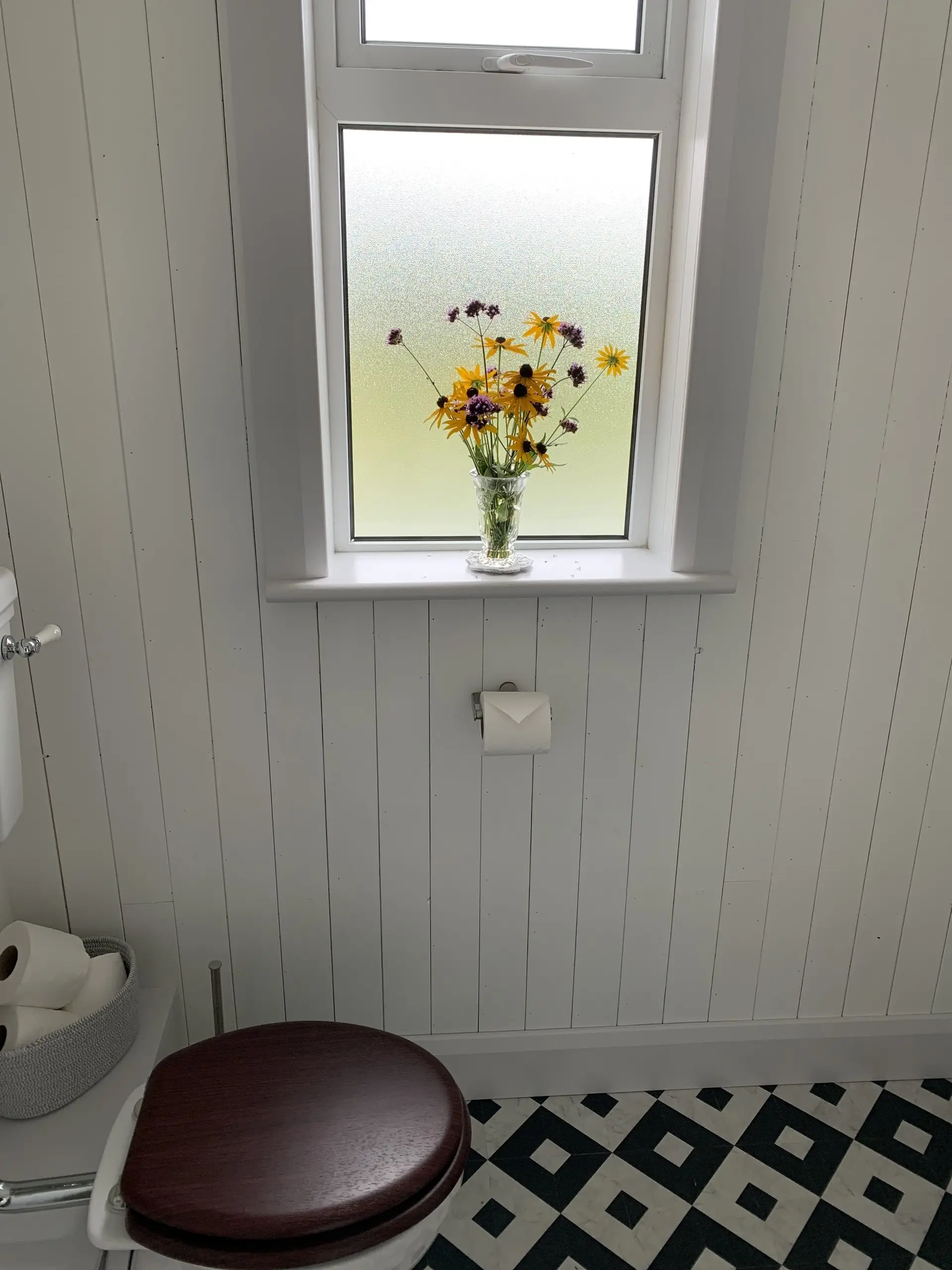 A walk-in shower with a glass screen, with fresh flowers on the windowsill and white panelled walls.