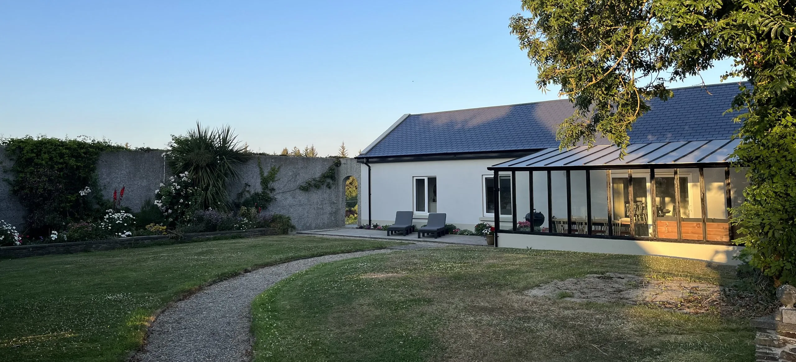 The Garden Cottage on a summer evening: a long white single-storey cottage with a slate roof and a glazed veranda lit warmly from inside, with sun loungers on the terrace and a gravel path curving across the lawn.