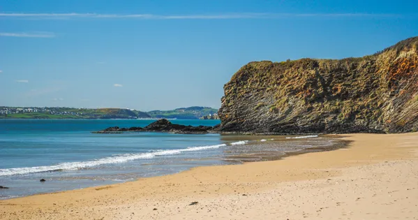 Dollar Bay: golden sand with small waves running in, beside a tall rocky outcrop, with the coastline visible across the water.