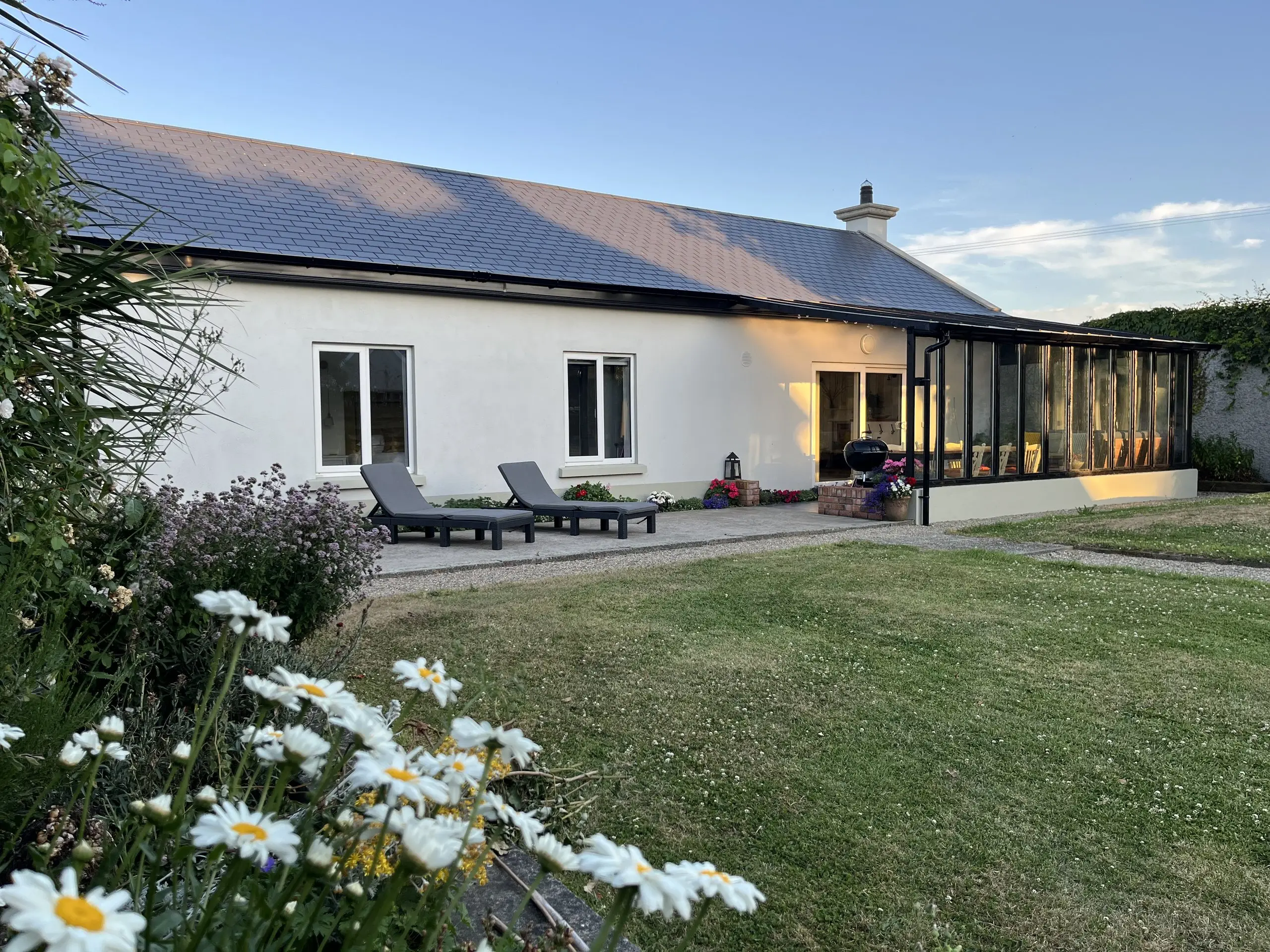 The cottage from the lawn with daisies in the foreground, sun loungers on the terrace and the veranda along the front.