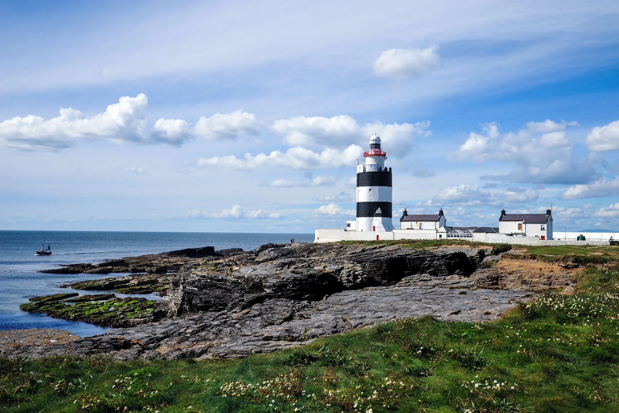 Hook Head lighthouse, banded black and white, standing above waves breaking on a rocky shore.