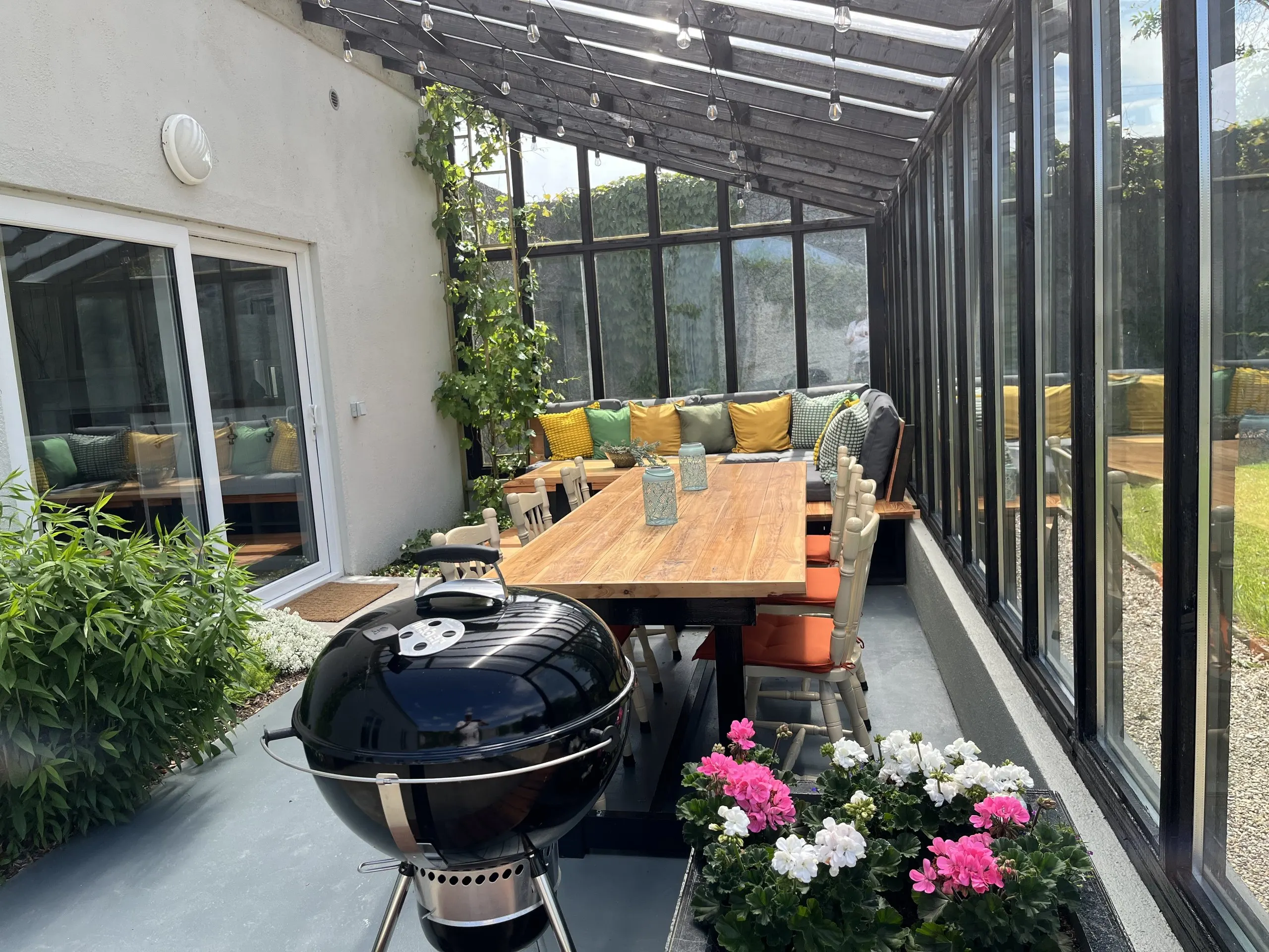The porch set for outdoor dining: a long oak table with orange-cushioned chairs, a kettle barbecue and flowers, under a glazed roof.