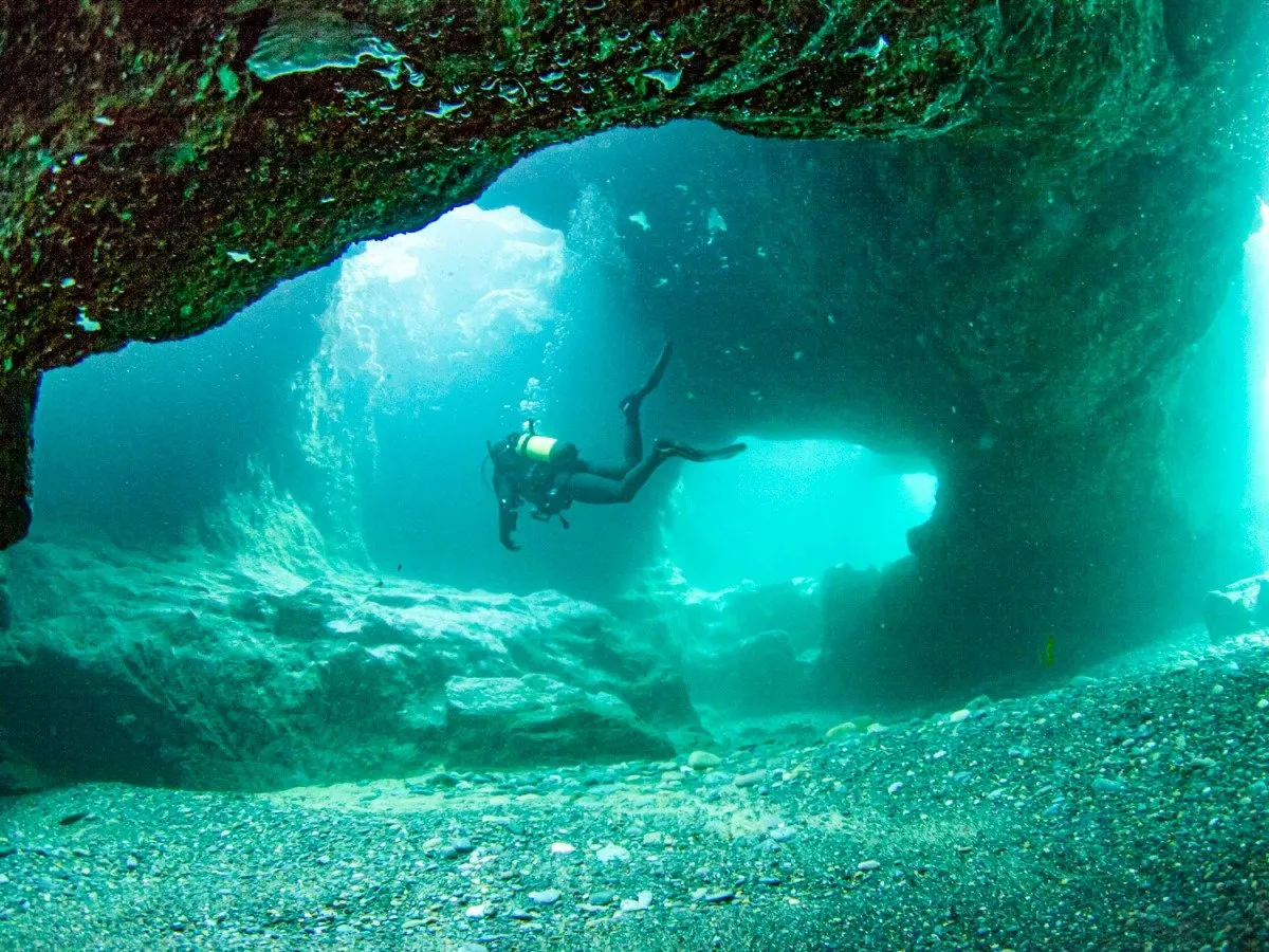 A scuba diver silhouetted in the mouth of an underwater cave at Hook Head, lit turquoise from the open sea behind.