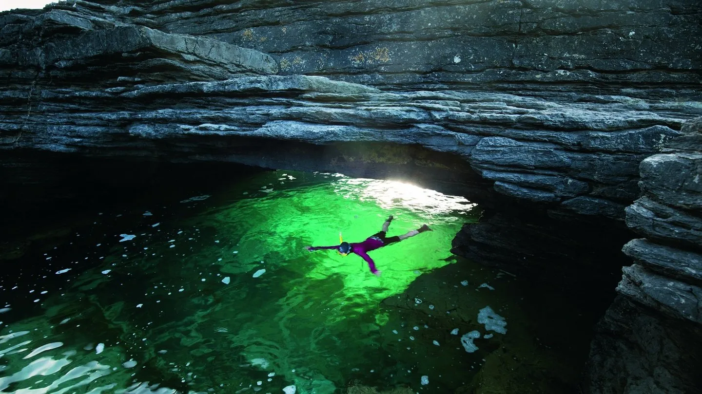 A snorkeller floating in a sea cave, the water lit vivid green from below.