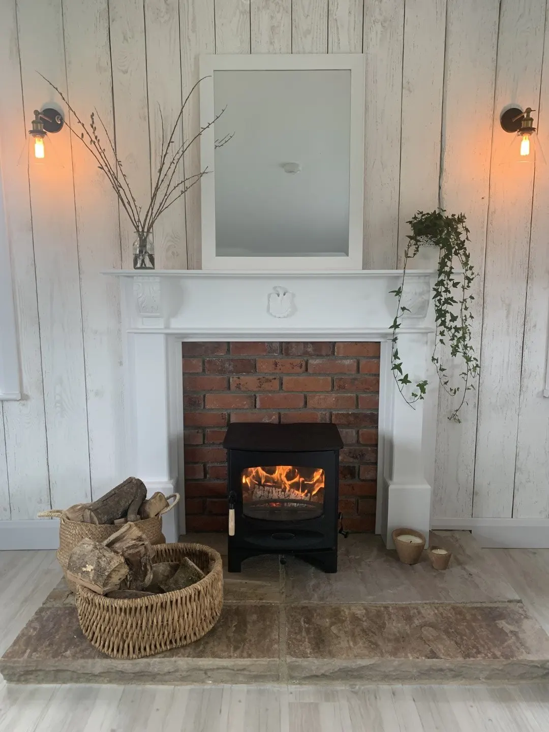 A lit wood-burning stove set in a brick fireplace with a white mantel and a basket of logs beside it.